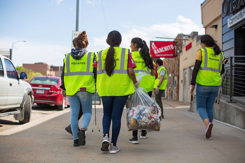 A group of neighborhood cleanup volunteers wearing bright yellow safety vests walking away from the camera. One volunteer is carrying a full bag of garbage.